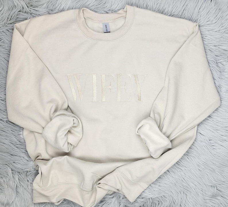 May include: A cream-colored sweatshirt with the word "WIFEY" embroidered in a light beige color.