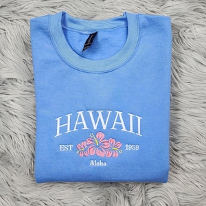 May include: A light blue sweatshirt with the text "HAWAII EST 1959 Aloha" and a pink embroidered hibiscus flower.