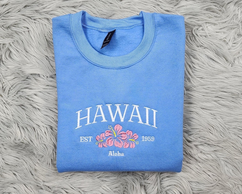 May include: A light blue sweatshirt with the text "HAWAII EST 1959 Aloha" and a pink embroidered hibiscus flower.