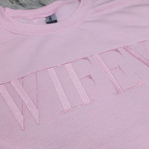 May include: A pink sweatshirt with the word "WIFEY" embroidered in white on the front.