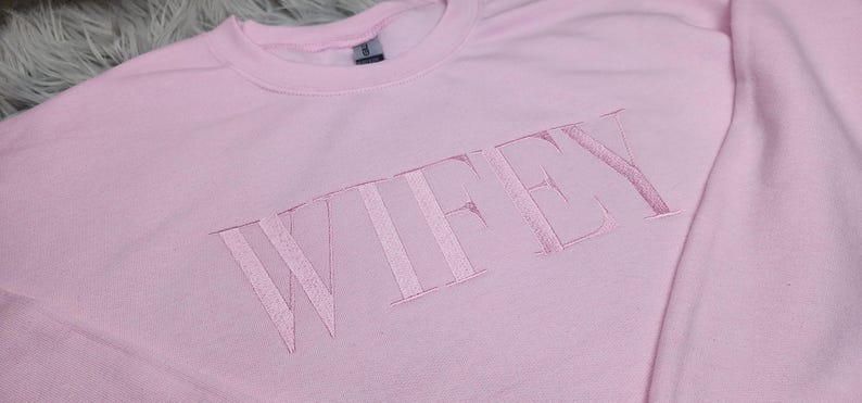 May include: A pink sweatshirt with the word "WIFEY" embroidered in white on the front.