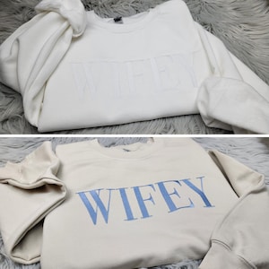May include: Two white sweatshirts with the word "WIFEY" embroidered on them. One sweatshirt has white embroidery and the other has light blue embroidery.
