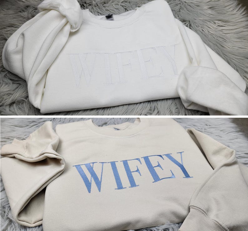 May include: Two white sweatshirts with the word "WIFEY" embroidered on them. One sweatshirt has white embroidery and the other has light blue embroidery.