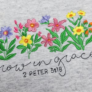 以下が含まれることがあります： グレーのトレーナーに刺繍された花と「grow in grace 2 Peter 3:18」の文字。