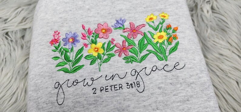 以下が含まれることがあります： グレーのトレーナーに刺繍された花と「grow in grace 2 Peter 3:18」の文字。