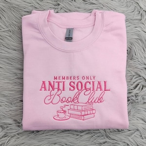May include: Pink sweatshirt with the text "Members Only Anti Social Book Club" embroidered in pink. The design includes a cup of coffee and a stack of books.