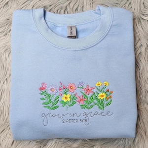 以下が含まれることがあります： 薄い青色のスウェットシャツに刺繍された花と「growing in grace 2 Peter 3:18」の文字