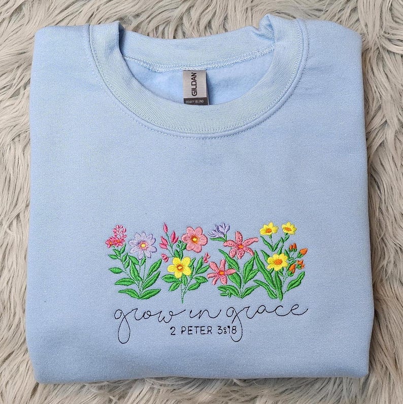 以下が含まれることがあります： 薄い青色のスウェットシャツに刺繍された花と「growing in grace 2 Peter 3:18」の文字