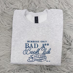 May include: Gray sweatshirt with the text "Members Only Bad A** Book Club Est. 29" in blue lettering. The text is stylized with a coffee cup and stack of books.