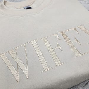 May include: A beige sweatshirt with the word "WIFEY" embroidered in a light beige color.