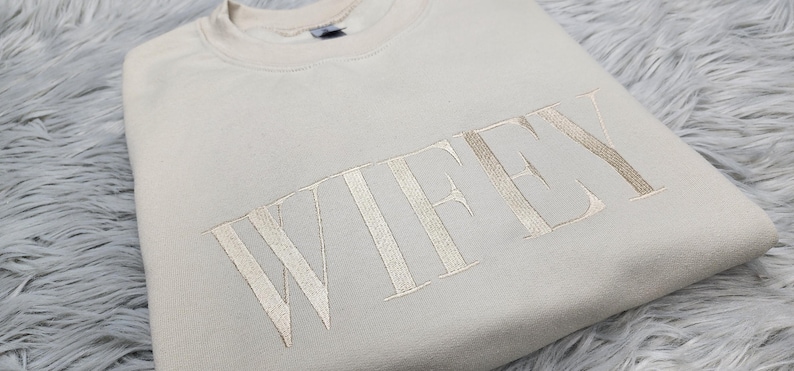 May include: A beige sweatshirt with the word "WIFEY" embroidered in a light beige color.