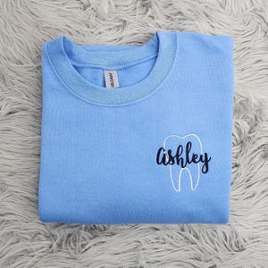 May include: Light blue sweatshirt with a white tooth graphic and the name "ashley" embroidered in black.