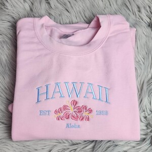 May include: Pink sweatshirt with embroidered text "HAWAII EST 1959" and a floral design with the word "Aloha" underneath.
