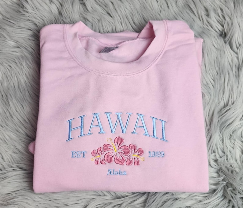 May include: Pink sweatshirt with embroidered text "HAWAII EST 1959" and a floral design with the word "Aloha" underneath.