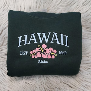 May include: A dark green sweatshirt with the text "HAWAII EST 1959" and the word "Aloha" printed on the front. The sweatshirt features a pink hibiscus flower design.