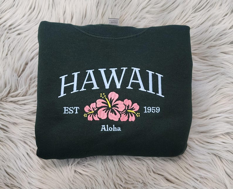 May include: A dark green sweatshirt with the text "HAWAII EST 1959" and the word "Aloha" printed on the front. The sweatshirt features a pink hibiscus flower design.