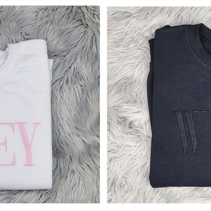May include: Two sweatshirts with the word "WIFEY" printed on them. One sweatshirt is white with pink lettering and the other is dark gray with black lettering.