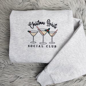 May include: A light gray sweatshirt with the embroidered text "Christmas Spirits Social Club." The design features three martini glasses with red and white candy cane straws and holly accents. A cozy, festive garment.