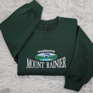 May include: A dark green sweatshirt with a white embroidered design featuring a mountain range and the text "WASHINGTON MOUNT RAINIER".