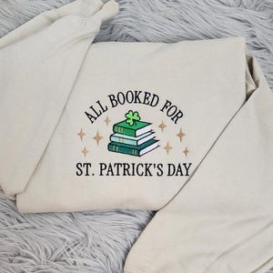 May include: A beige sweatshirt with the text "All Booked for St. Patrick's Day" embroidered on the front. The text is surrounded by gold stars and a stack of books with a shamrock on top.