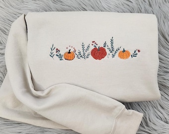 May include: A cream-colored sweatshirt with an embroidered design of pumpkins and leaves in shades of orange, green, and red.