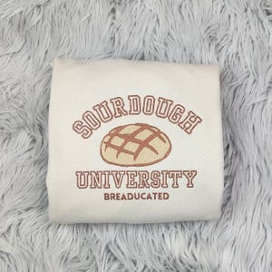 May include: A white sweatshirt with the text "Sourdough University" and the words "BREADUCATED" underneath. A brown bread roll with a crosshatch pattern is centered above the text.
