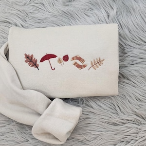 May include: A white sweatshirt with embroidered autumn-themed imagery, including leaves, an umbrella, and acorns.