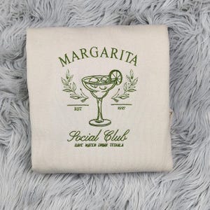 Pu&ograve; includere: Una borsa di tela beige con ricamo verde di un bicchiere di margarita, uno spicchio di lime e il testo "Margarita Social Club Est. 1997 Save Water Drink Tequila".