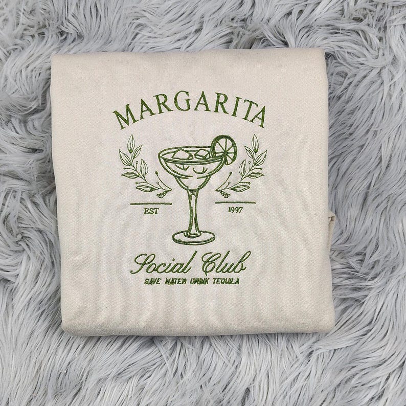 Pu&ograve; includere: Una borsa di tela beige con ricamo verde di un bicchiere di margarita, uno spicchio di lime e il testo "Margarita Social Club Est. 1997 Save Water Drink Tequila".