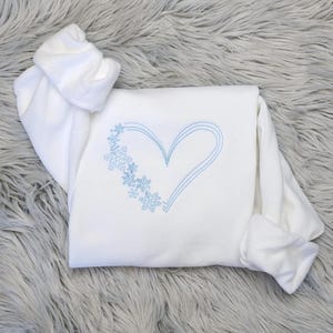 Pu&ograve; includere: Felpa bianca con un design a cuore e fiocchi di neve ricamati in blu. La parte superiore a maniche lunghe &egrave; piegata su una superficie grigia e soffice. Il cuore &egrave; delineato da tre linee parallele.