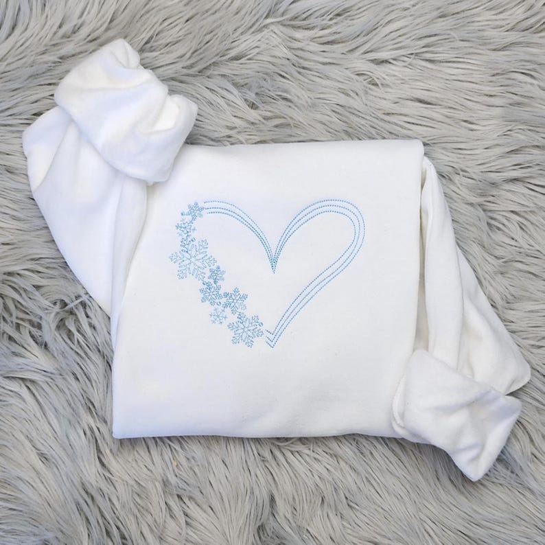 Pu&ograve; includere: Felpa bianca con un design a cuore e fiocchi di neve ricamati in blu. La parte superiore a maniche lunghe &egrave; piegata su una superficie grigia e soffice. Il cuore &egrave; delineato da tre linee parallele.