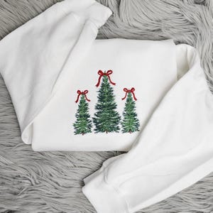 May include: White sweatshirt featuring three embroidered Christmas trees with red bows. The trees are green with detailed needlework, and the bows add a festive touch. The sweatshirt is folded, showcasing the design prominently.