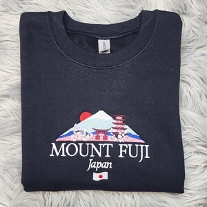 Może przedstawiać: Czarna bluza z haftowanym wzorem g&oacute;ry Fudżi, z czerwonym słońcem, pagodą i napisem "MOUNT FUJI Japan". Projekt zawiera japońską flagę. Bluza jest złożona na białej, puszystej powierzchni.