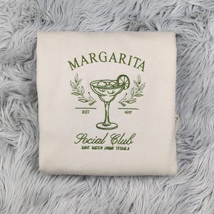 Pu&ograve; includere: Una felpa bianca con collo a girocollo con testo ricamato in verde che recita "Margarita Social Club Est. 1997 Save Water Drink Tequila". Il design include anche un bicchiere di margarita ricamato in verde con una fetta di lime.