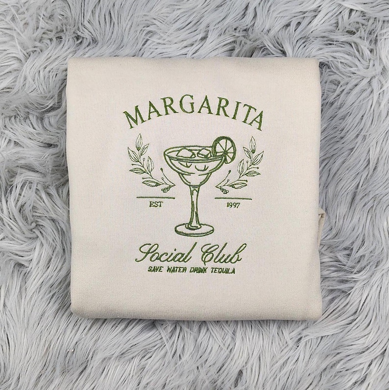 Pu&ograve; includere: Una felpa bianca con collo a girocollo con testo ricamato in verde che recita "Margarita Social Club Est. 1997 Save Water Drink Tequila". Il design include anche un bicchiere di margarita ricamato in verde con una fetta di lime.