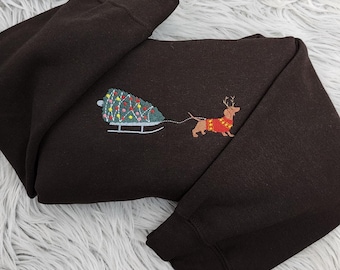 Puede incluir: Una sudadera marrón oscuro con un diseño navideño bordado. El diseño presenta un perro salchicha con un suéter rojo y cuernos tirando de un trineo con un árbol de Navidad decorado. La sudadera está doblada sobre una superficie blanca y esponjosa.