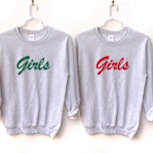 以下が含まれることがあります： 「Girls」とプリントされたグレーのトレーナーが2枚。1枚は緑色の文字で、もう1枚は赤色の文字で書かれています。