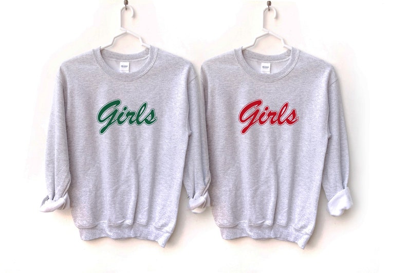 以下が含まれることがあります： 「Girls」とプリントされたグレーのトレーナーが2枚。1枚は緑色の文字で、もう1枚は赤色の文字で書かれています。