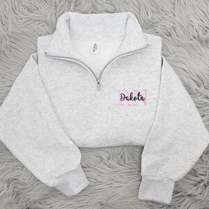 May include: Gray quarter-zip sweatshirt with pink embroidered letters "RN" and the name "Dakota" below.