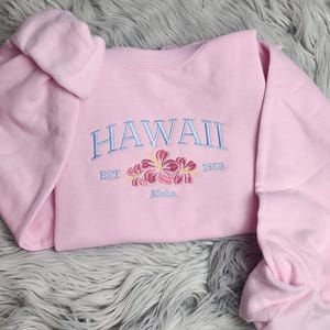 May include: Pink sweatshirt with the text "HAWAII EST 1953 Maka" embroidered in blue with pink flowers.