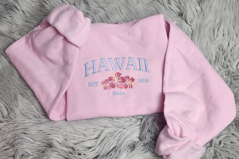 May include: Pink sweatshirt with the text "HAWAII EST 1953 Maka" embroidered in blue with pink flowers.
