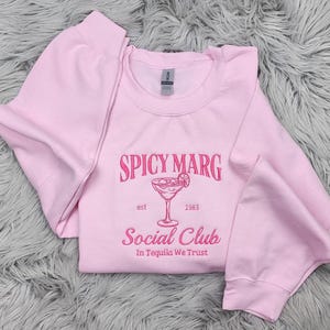 May include: Pink crewneck sweatshirt with the text "Spicy Marg Social Club est 1983 In Tequila We Trust" and a pink illustration of a margarita glass with a lime wedge.