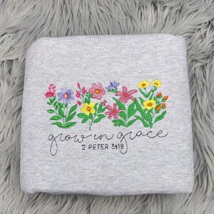 以下が含まれることがあります： グレーのトレーナーに刺繍された花と「grow in grace 2 Peter 3:18」の文字。