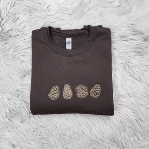 May include: A brown sweatshirt with a design of four pine cones in a row on the front.
