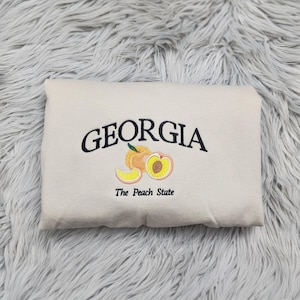 May include: A cream colored sweatshirt with the text "GEORGIA The Peach State" embroidered in black. The design includes a peach with a slice of peach next to it.