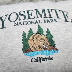 May include: Gray sweatshirt with an embroidered Yosemite National Park logo featuring a bear, trees, and a river. The text "Yosemite National Park" and "California" are embroidered in green.