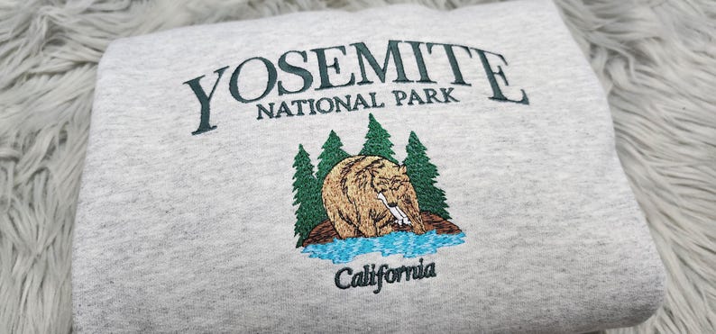 May include: Gray sweatshirt with an embroidered Yosemite National Park logo featuring a bear, trees, and a river. The text "Yosemite National Park" and "California" are embroidered in green.