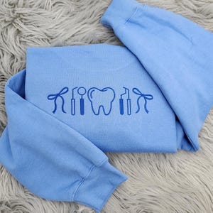 May include: A light blue sweatshirt with a dental-themed design. The design features a tooth, dental tools, and a bow. The sweatshirt is folded and placed on a fluffy, gray surface.
