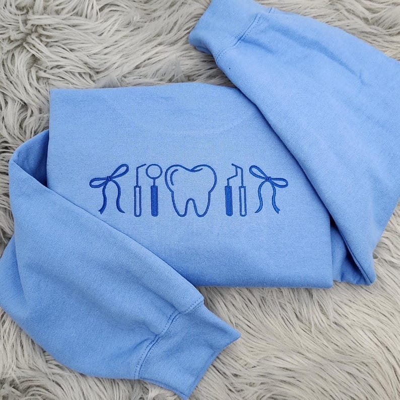 May include: A light blue sweatshirt with a dental-themed design. The design features a tooth, dental tools, and a bow. The sweatshirt is folded and placed on a fluffy, gray surface.