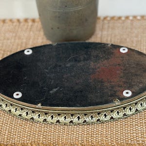 May include: An oval-shaped vanity tray featuring a dark, weathered backing with four white circular feet. The tray is enclosed by a decorative, perforated gold-toned metal rim with a repeating leaf-like pattern along the entire outer edge.